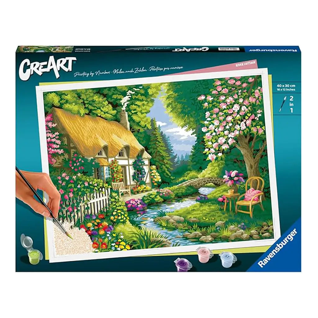 Ravensburger CreArt paint by numbers kit box showing River Cottage scene with thatched roof house, lush garden, flowering trees, and a winding stream in vivid colors.