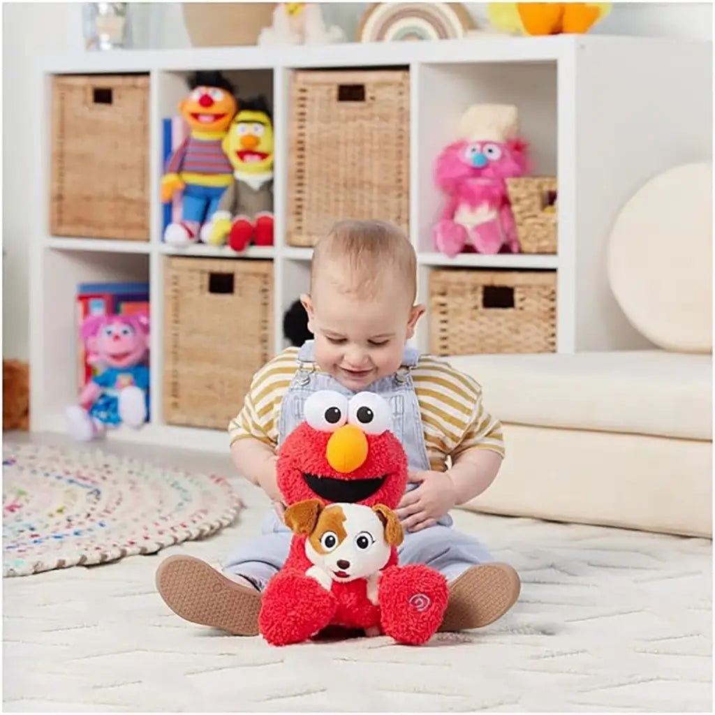 Baby seated on playroom floor with plush Elmo holding puppy Tango, colorful cubbies with Sesame Street toys behind, showing toy’s size and interactive appeal for toddlers.