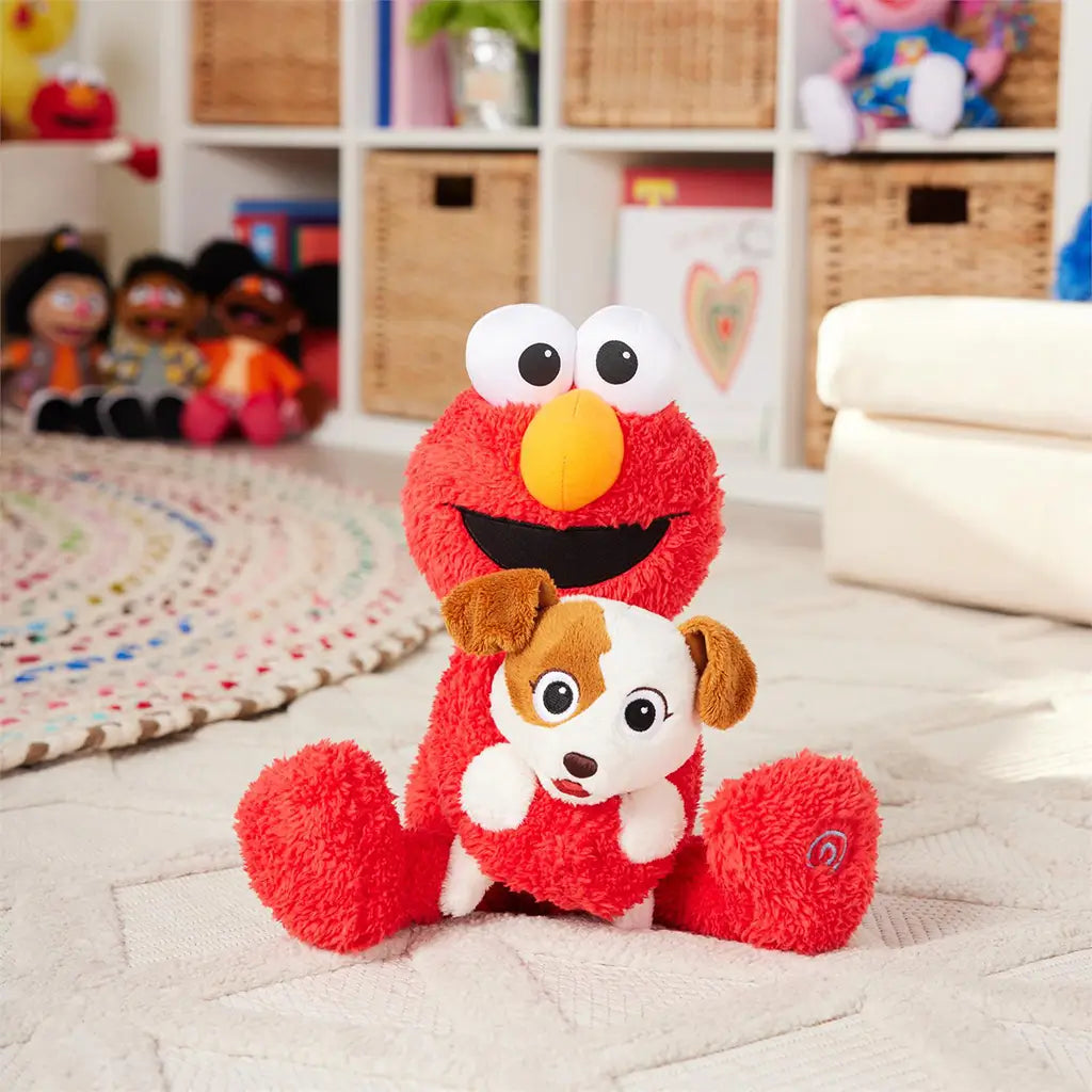 Plush Elmo holding puppy Tango toy sitting on white rug in playroom with wicker storage cubbies and Sesame Street dolls, staged to highlight toy’s interactive design.