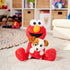 Plush Elmo holding puppy Tango toy sitting on white rug in playroom with wicker storage cubbies and Sesame Street dolls, staged to highlight toy’s interactive design.