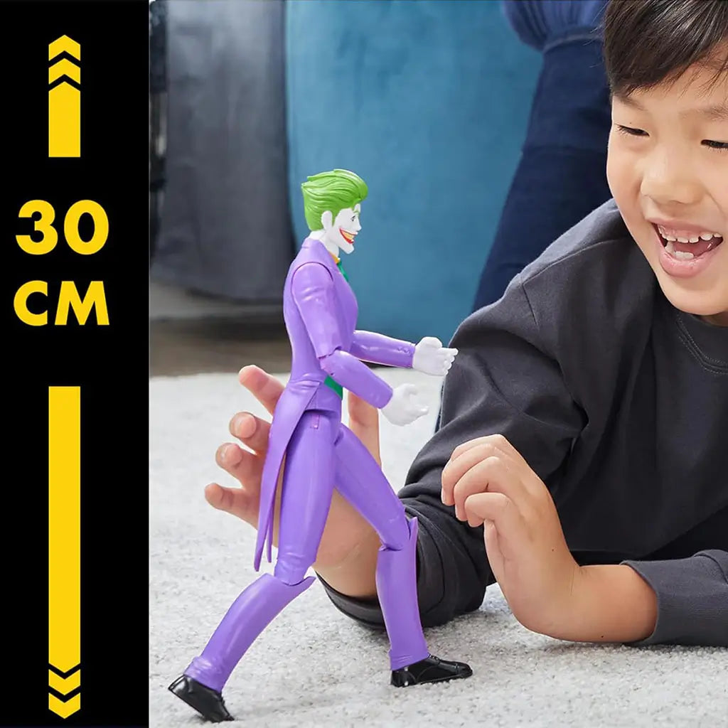 Joker 12-inch DC Comics action figure shown beside 30 cm height graphic, featuring purple tailcoat, white gloves, and green hair while a child plays in background.