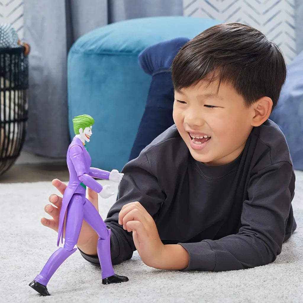 Smiling boy plays on floor with DC Comics Joker 12-inch action figure, posed walking forward in purple suit, showing articulated limbs for creative play.