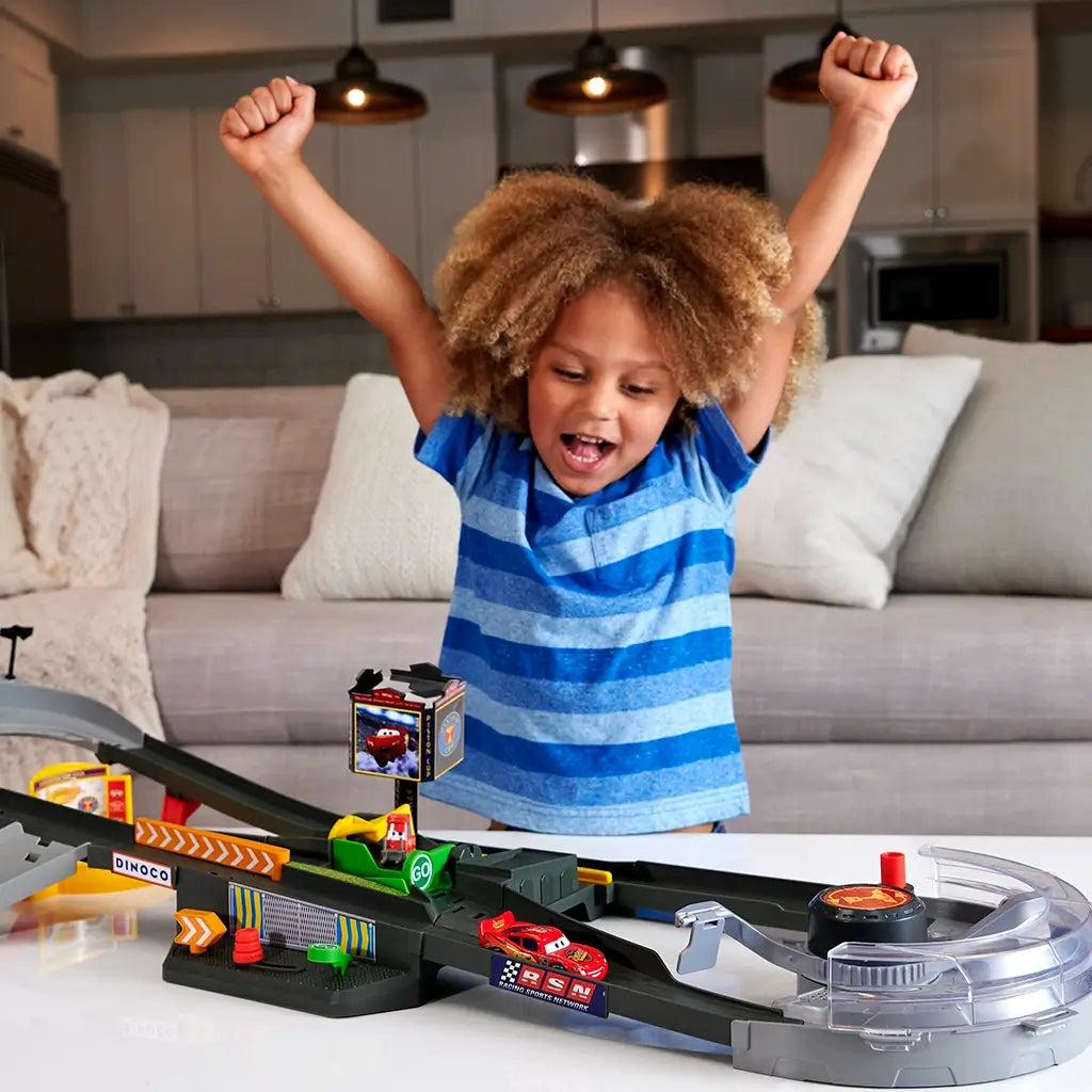 Disney Pixar Cars toy playset shown with a young child raising arms in excitement, featuring a dark gray plastic race track, yellow ramps, and Lightning McQueen car on a tabletop in a bright living room setting.