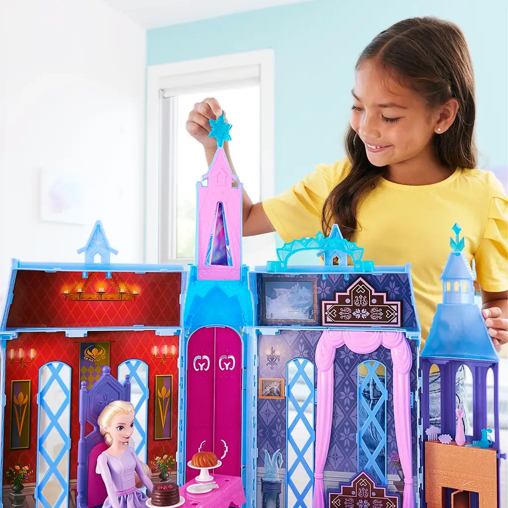 Smiling child in yellow shirt playing with Frozen Arendelle Castle playset, arranging Elsa doll, furniture, and transforming pieces inside the colorful fold-out castle.