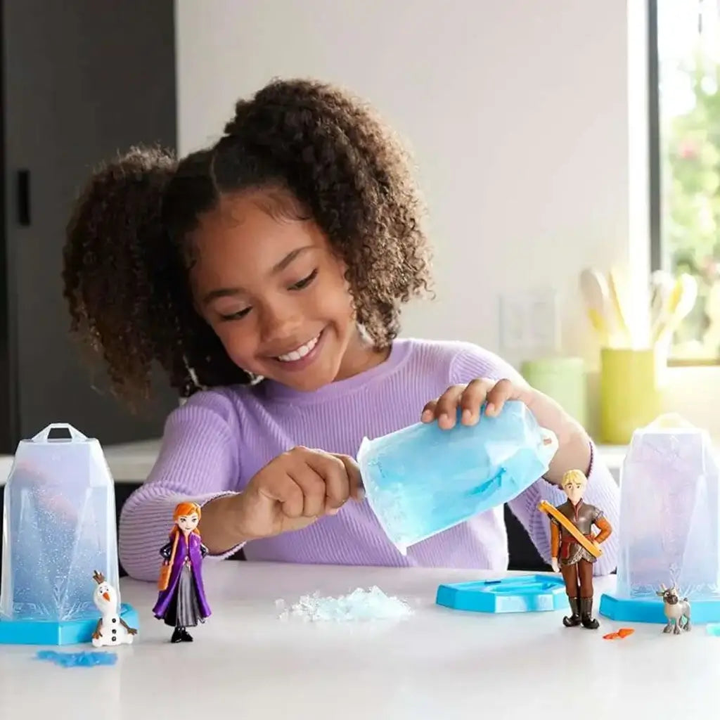 Smiling child playing with Disney Frozen Ice Reveal figures of Anna, Kristoff, and Olaf while scooping blue ice gel from a crystal-shaped container in a bright, cozy room setting.