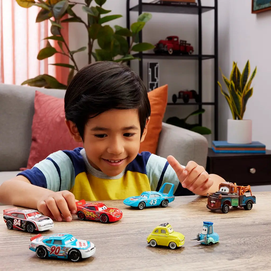 Smiling young boy in striped shirt playing at a wooden table with multiple Disney Pixar Cars diecast toy vehicles including Lightning McQueen and Acer, indoors near plants.