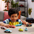 Smiling young boy in striped shirt playing at a wooden table with multiple Disney Pixar Cars diecast toy vehicles including Lightning McQueen and Acer, indoors near plants.