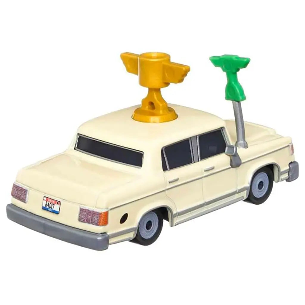 Disney Pixar Cars Brad Winner diecast shown from a rear angled view, highlighting the cream metal body, squared trunk and bumper, gold roof trophy mount, green microphone arm, gray wheels, and classic sedan proportions on white.
