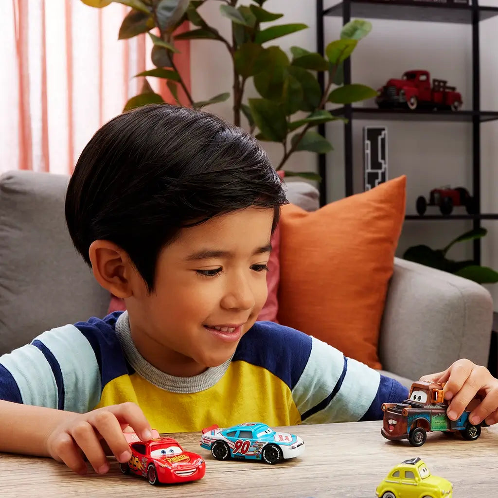 Disney Pixar Cars diecast vehicles in a lifestyle scene with a smiling child playing at a table, colorful metal character cars, rounded toy shapes, and a warm living room background.