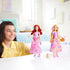 A smiling girl plays with two Disney Princess fashion dolls—Ariel and Rapunzel—both wearing floral skirts and pastel tops in a bright, cozy bedroom setting. The Ariel doll holds a fork accessory, while the Rapunzel doll carries a woven basket with a flower, surrounded by small toy pastries on a white table.