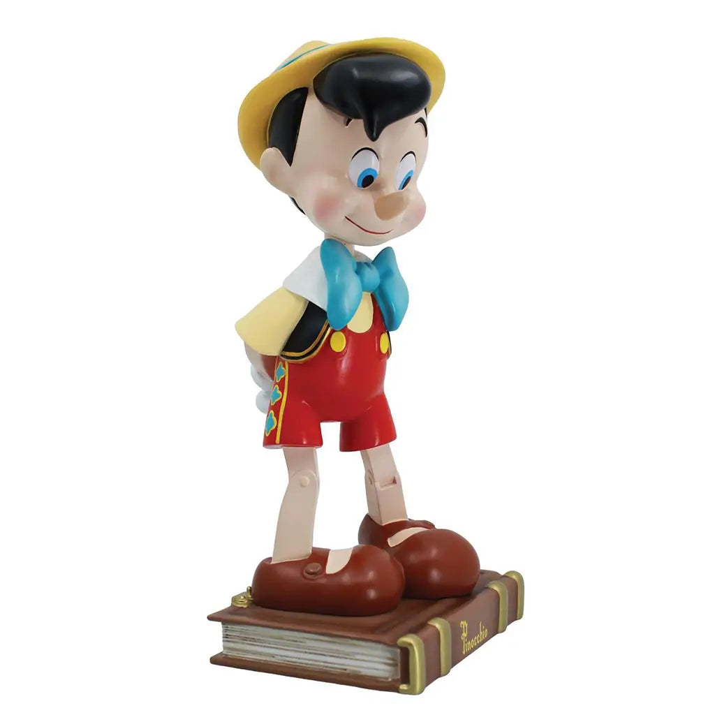 Angled front view of Disney Pinocchio figure with big eyes and shy expression, wearing red outfit and standing on a brown book base; 85th anniversary collectible.
