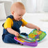 A baby in denim overalls plays with the Fisher-Price Storybook Rhymes toy, turning colorful plastic pages that display friendly animal illustrations in a bright, soft-lit home setting.