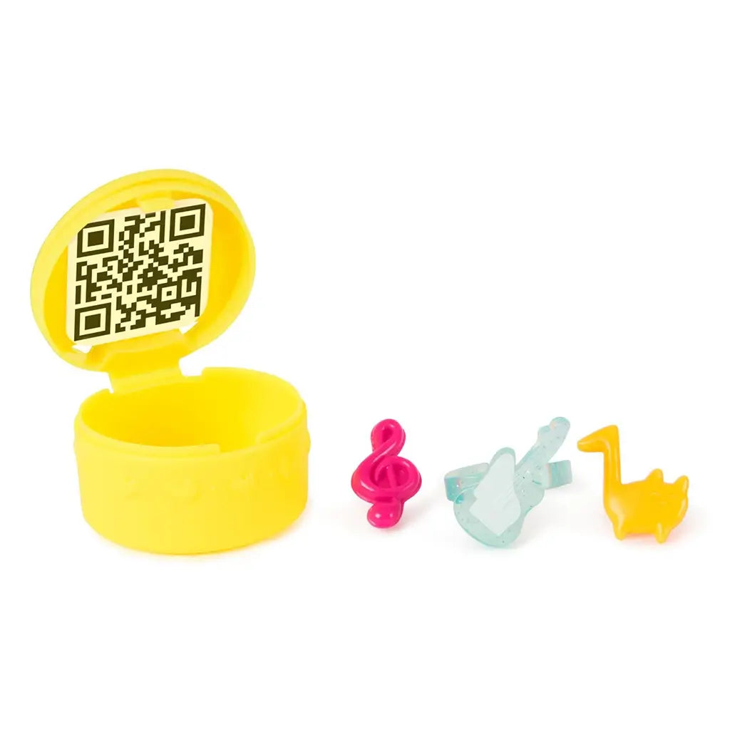 Gabby's Dollhouse surprise accessory capsule in glossy yellow plastic with hinged lid revealing QR code inside, shown open beside small music-themed toy pieces on a white surface.