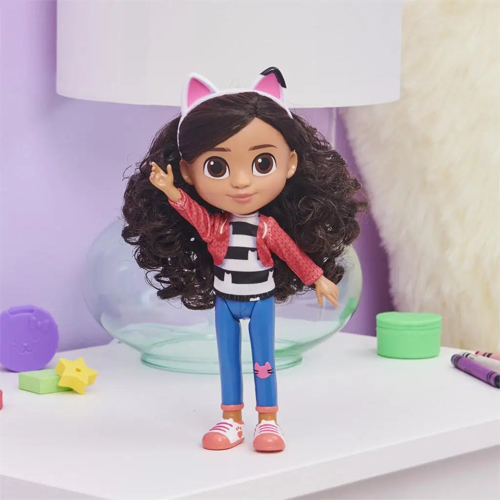 Gabby's Dollhouse Gabby Girl doll in coral pink jacket and fitted blue jeans, molded plastic fashion figure with voluminous curly hair and white cat-ear headband, posed with arm raised on a bedroom desk.