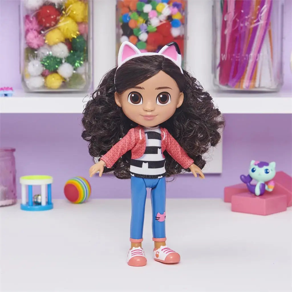 Gabby's Dollhouse Gabby Girl doll in pink jacket, black-and-white striped shirt, and blue jeans with cat patch, smooth plastic figure with thick curly dark hair and cat-ear headband, standing in a pastel craft room.