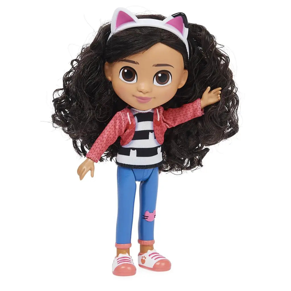 Gabby's Dollhouse Gabby Girl doll wearing textured pink jacket, striped top, and blue jeans, smooth plastic fashion figure with large brown eyes and full curly hair, waving against a white studio background.