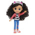 Gabby's Dollhouse Gabby Girl doll wearing textured pink jacket, striped top, and blue jeans, smooth plastic fashion figure with large brown eyes and full curly hair, waving against a white studio background.