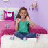 Smiling young girl sitting on a white rug in a purple bedroom holding two Gabby’s Dollhouse mini figures, surrounded by toy capsules and packaging from the DreamWorks Netflix series.