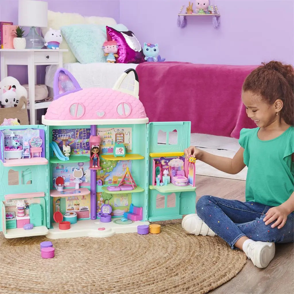 Gabby’s Dollhouse playset in pastel plastic colors with multiroom dollhouse structure and small figurines, child playing on floor beside detailed rooms, soft bedroom setting with toys and decor in background.