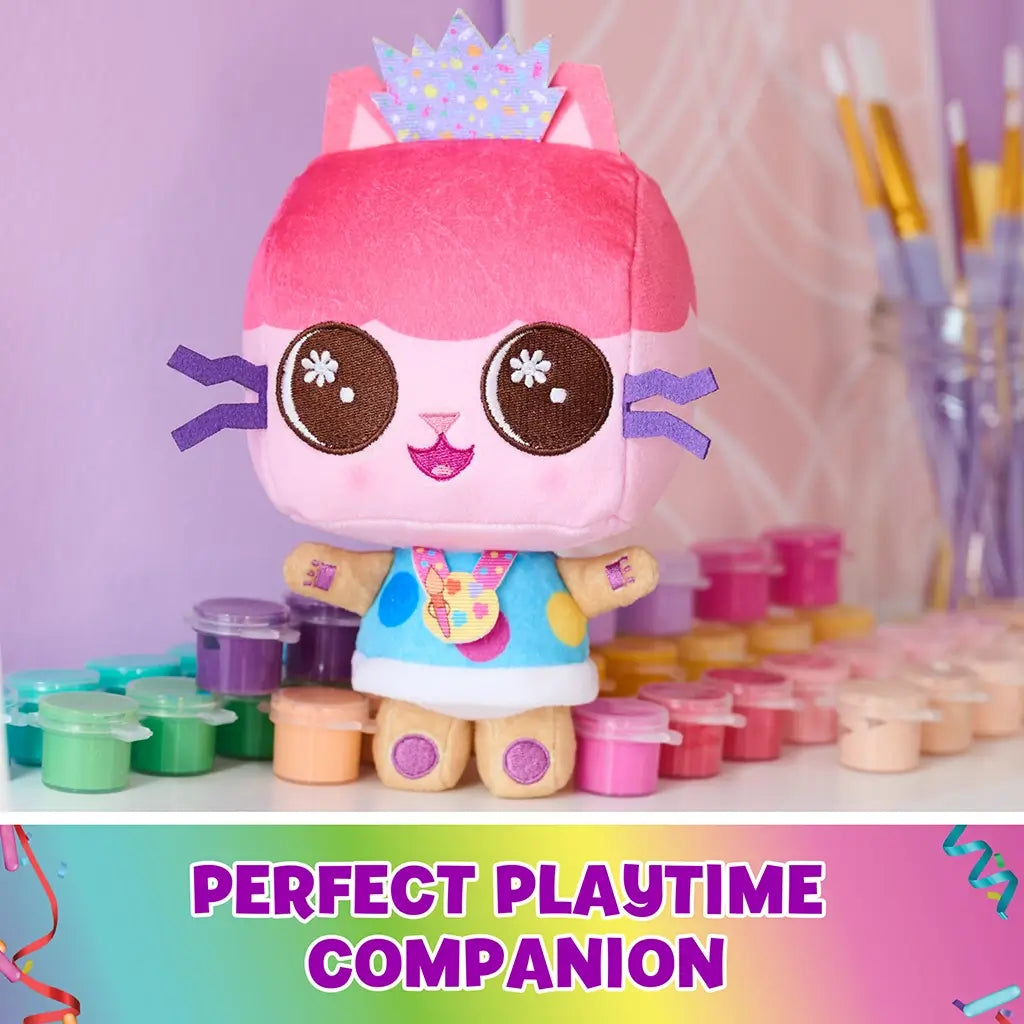 Gabby’s Dollhouse plush toy featuring pink soft textured fabric in cube-shaped cat design with embroidered eyes and crown, standing on a table surrounded by paint pots in a bright creative playroom setting.