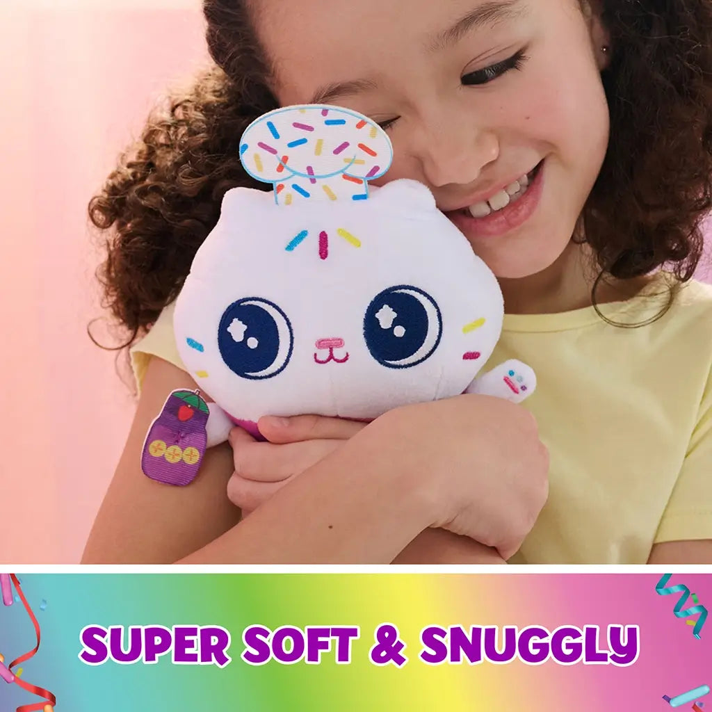 Gabby’s Dollhouse Cakey Cat plush toy white soft textured cupcake cat with embroidered face, sprinkle accents and rainbow base, cuddly character hugged by child in cozy bedroom scene.