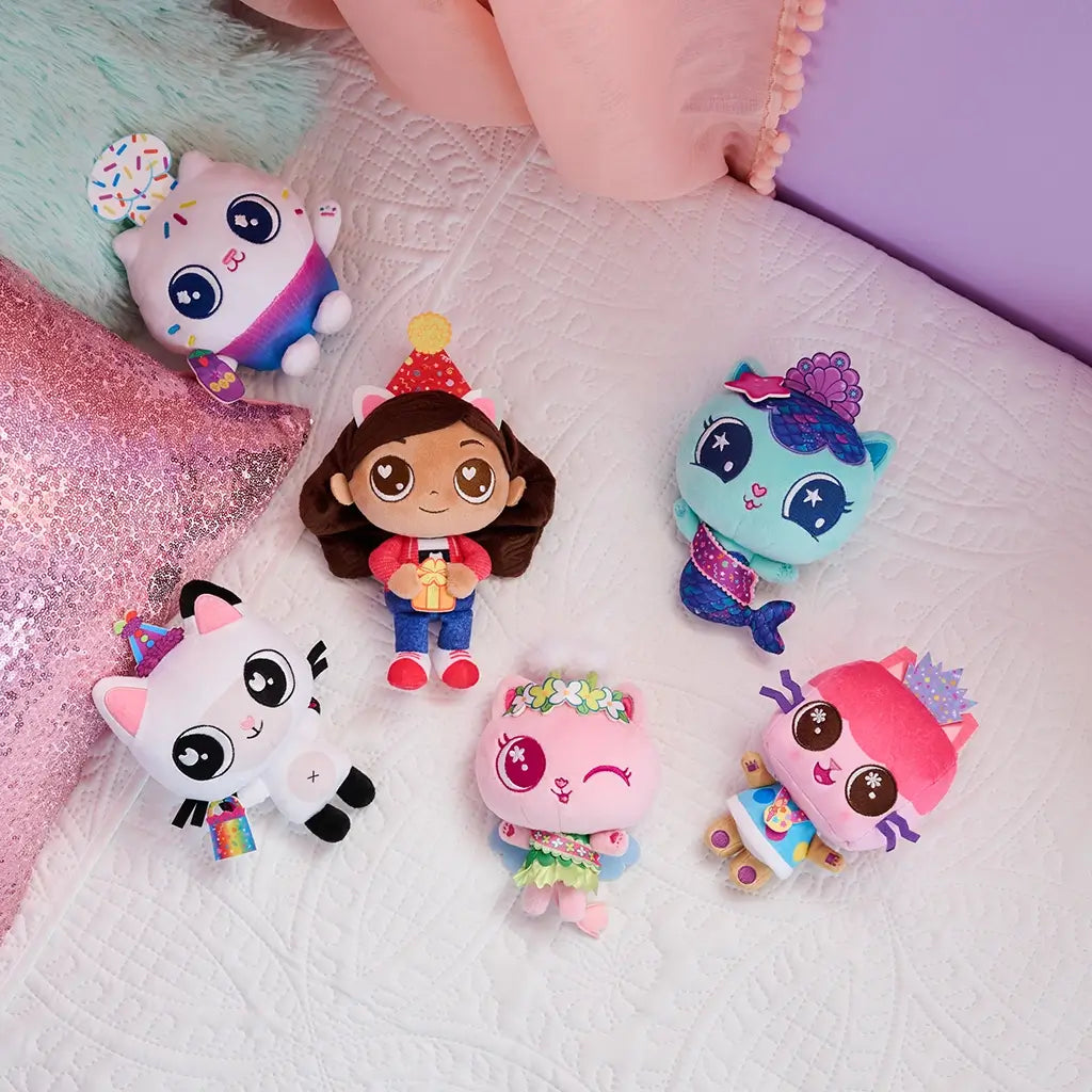 Gabby's Dollhouse plush toys in bright pastel fabrics, small rounded character shapes with embroidered faces, arranged on a quilted bed, showcasing collectible set and playful themed characters.