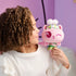 Gabby's Dollhouse Cakey Cat plush toy in pastel pink fabric, small rounded kitten shape with stitched face and floral crown, held by a smiling child in a cozy bedroom, highlighting soft cuddly play.