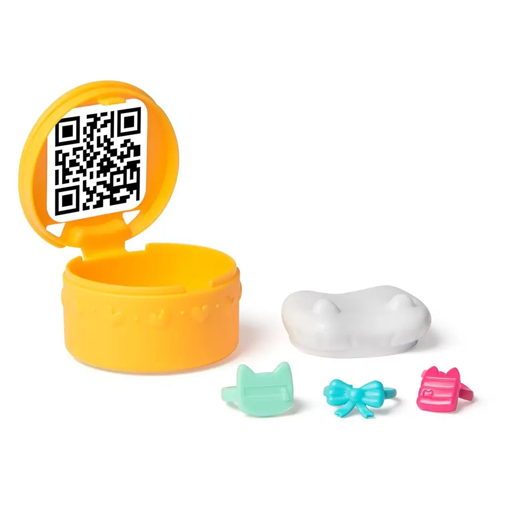 Gabby's Dollhouse surprise accessory capsule in glossy yellow plastic with hinged lid showing QR code inside, displayed open beside tiny costume pieces on a white surface.