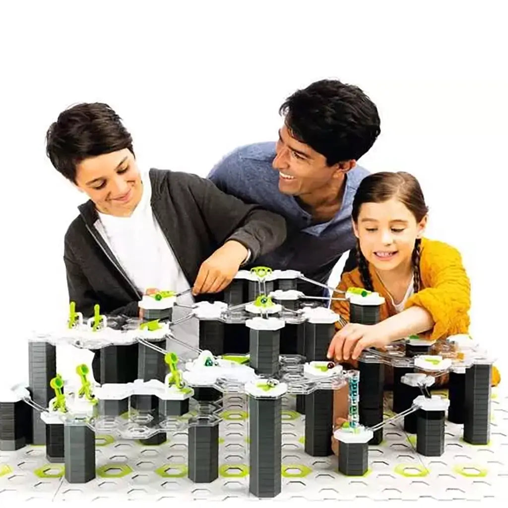 Smiling father with two children builds a multi-level GraviTrax marble run using white hexagonal tiles, black towers, and green action elements on a large base grid.