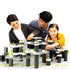 Smiling father with two children builds a multi-level GraviTrax marble run using white hexagonal tiles, black towers, and green action elements on a large base grid.