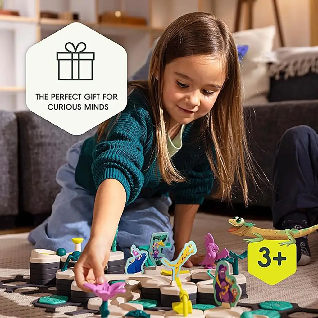 Young girl smiling while arranging pink and green desert-themed figures on GraviTrax Junior tiles, with banner noting the set as a perfect gift for curious kids ages three and up.