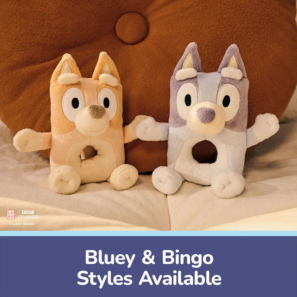 Bluey and Bingo plush ring rattles feature pastel blue and tan soft fabric, rounded blocky shapes, embroidered faces, and open center rings, offering gentle tactile play for babies on soft bedding.