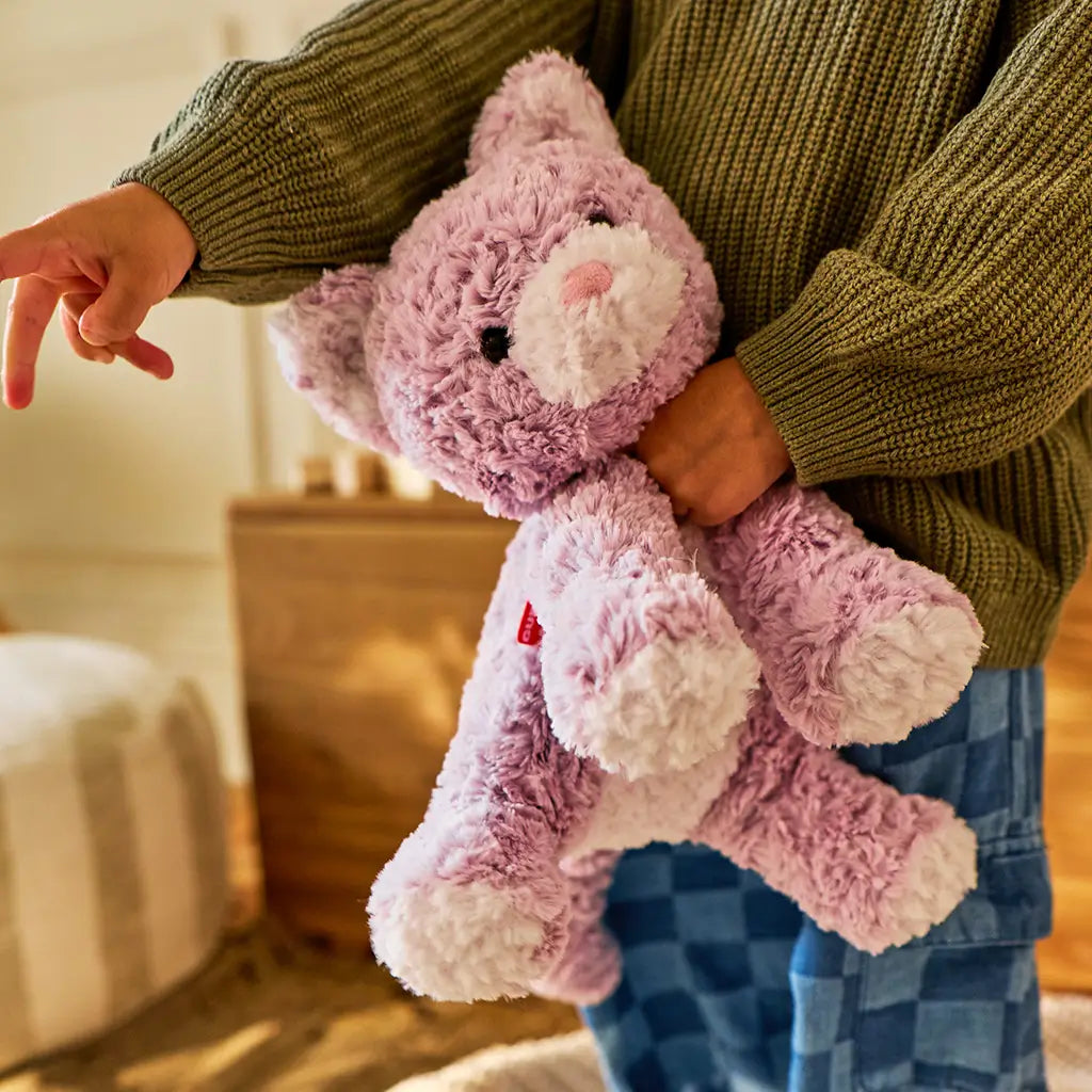 GUND Cozys Kitten stuffed animal in light purple plush fabric with rounded limbs and floppy posture, positioned upright on a soft rug near wooden furniture to show comforting size and cozy home setting.