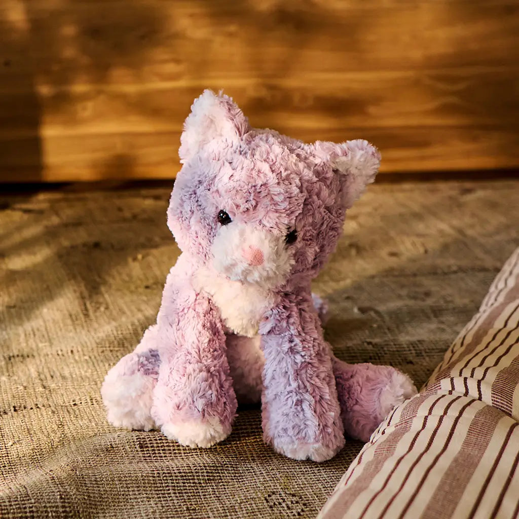 GUND Cozys Kitten plush toy in pastel purple polyester fur with a fuzzy surface and relaxed seated form, displayed on a textured rug indoors to highlight cozy décor appeal and everyday playtime companionship.