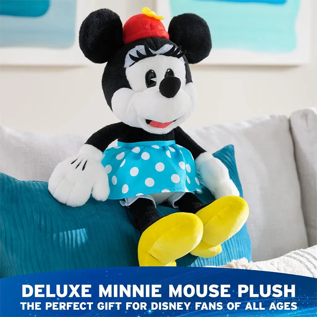 Disney Minnie Mouse plush in black and white soft fabric with rounded seated character form, red hat with yellow flower, blue polka dot skirt, white gloves, and yellow shoes, staged on a couch as a deluxe plush gift for display or play.