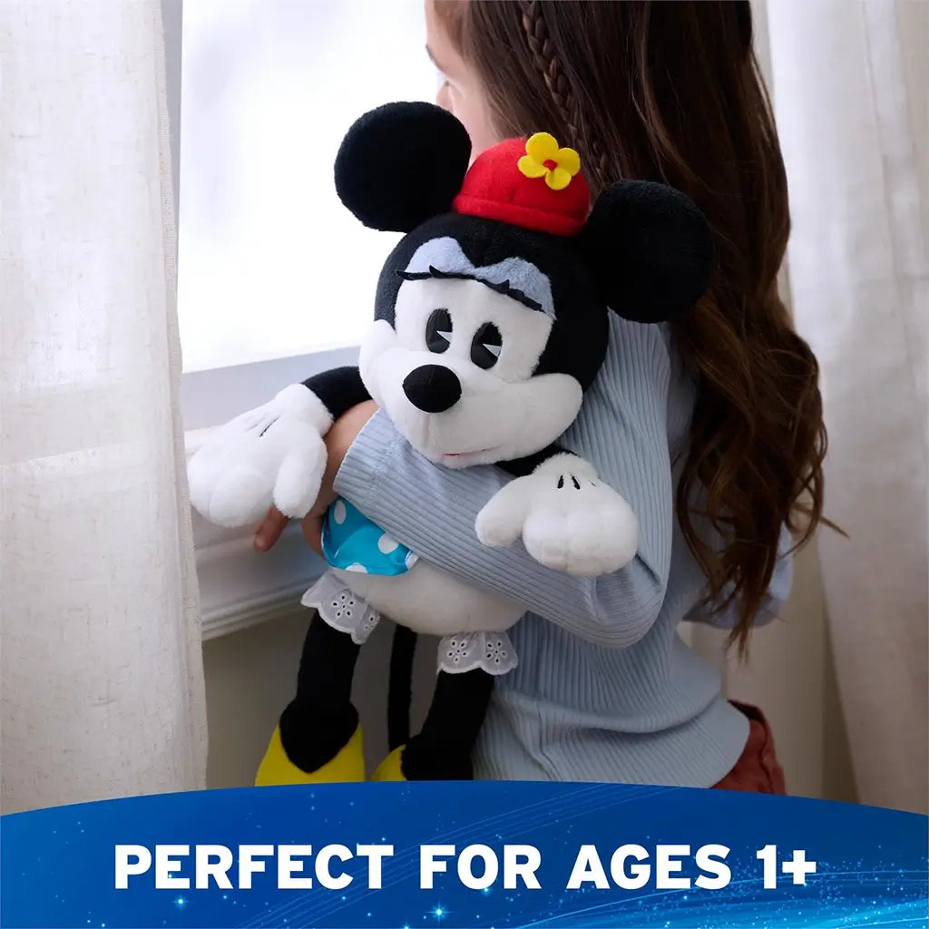 Disney Minnie Mouse plush toy in black and white soft fabric with seated character shape, red hat with yellow flower, blue polka dot skirt, white gloves, and yellow shoes, shown hugged by a child on a sofa to highlight suitability for ages one plus.