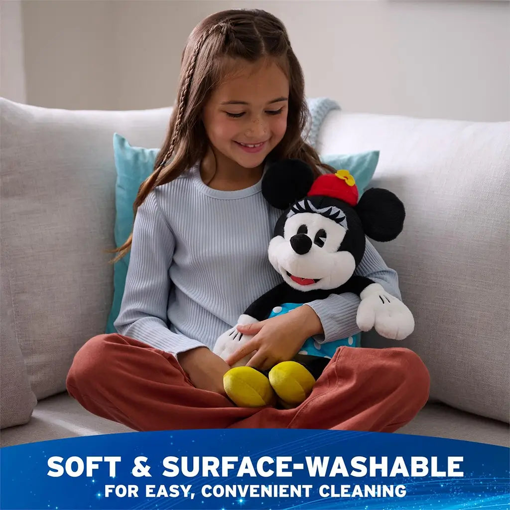 Disney Minnie Mouse plush in black and white fuzzy fabric with seated character shape, red hat with yellow flower, blue polka dot skirt, white gloves, and yellow shoes, shown with a child indoors to emphasize soft surface washable and easy care.