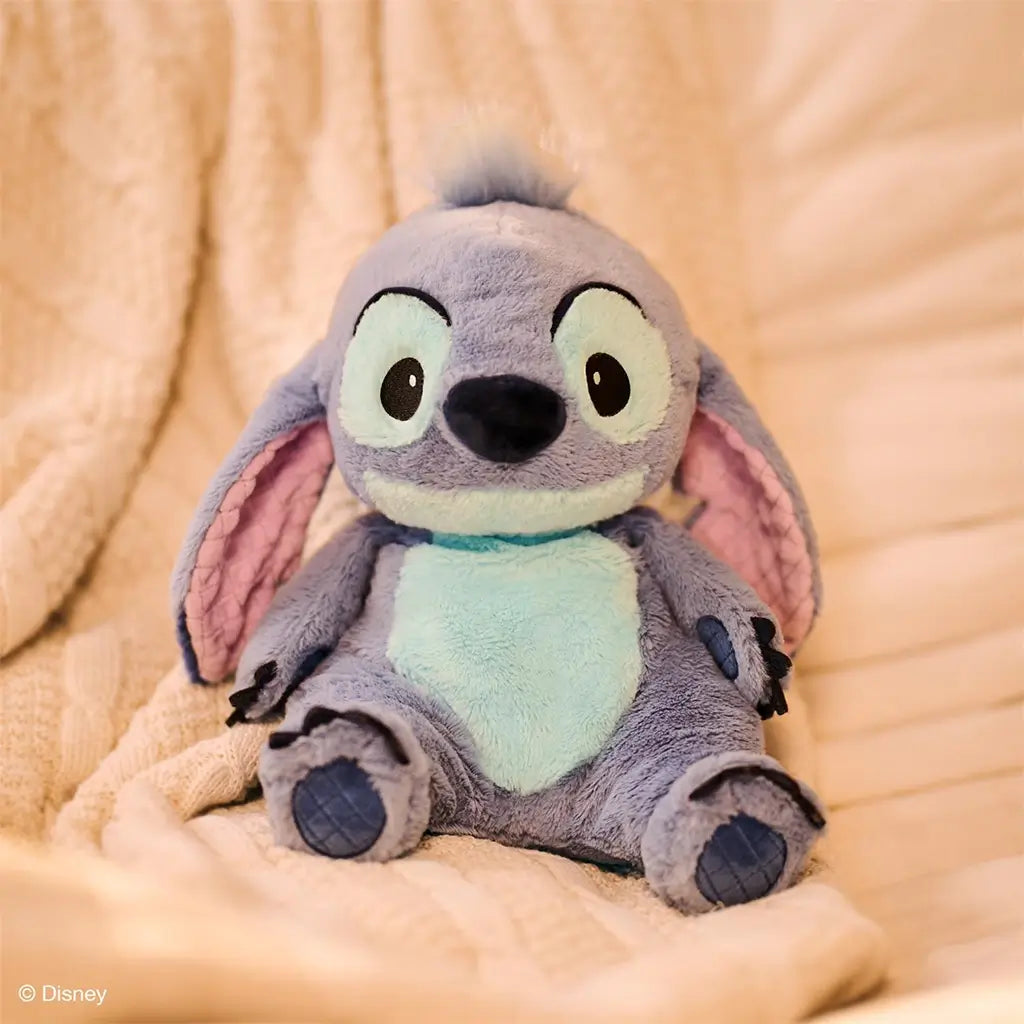 Disney Stitch plush toy in soft blue and lavender fabric with velvety texture, rounded ears, and embroidered eyes, designed for cuddling, seated on a cream quilted blanket in a cozy indoor setting.