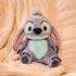 Disney Stitch plush toy in soft blue and lavender fabric with velvety texture, rounded ears, and embroidered eyes, designed for cuddling, seated on a cream quilted blanket in a cozy indoor setting.