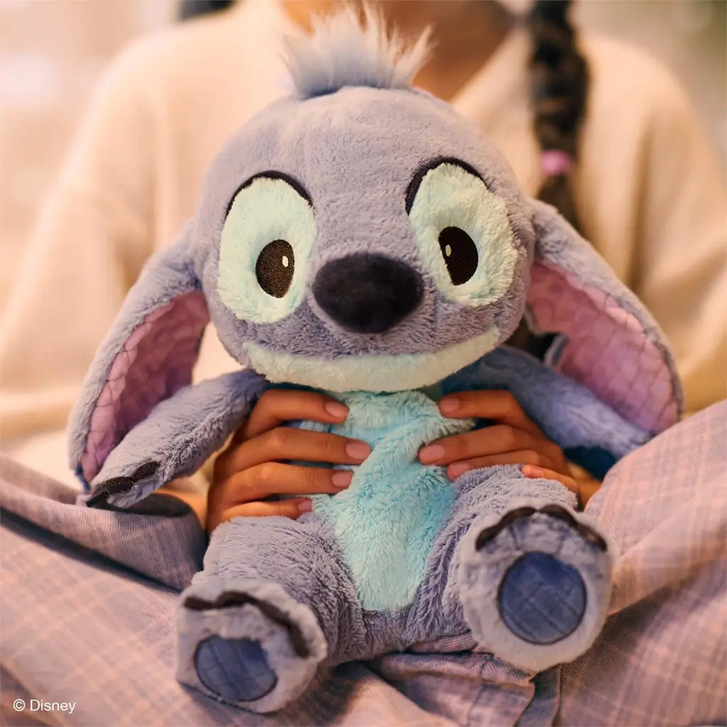 Disney Stitch plush toy crafted from ultra-soft blue fabric with floppy ears and padded feet, intended for cuddling, resting in a child’s hands while seated comfortably on a bed indoors.