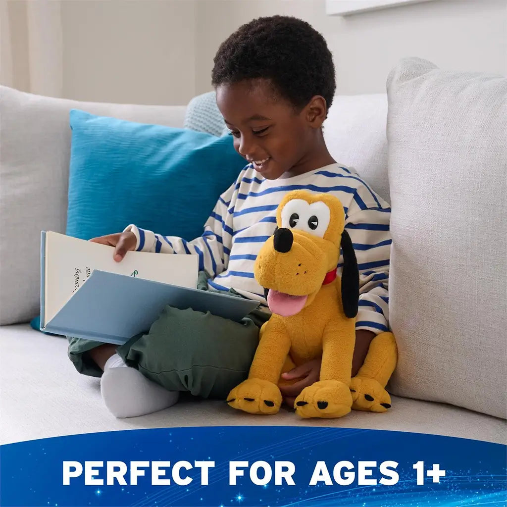 Disney Pluto plush in golden yellow soft fabric with rounded seated dog form, floppy black ears, pink tongue, and red collar, shown beside a smiling child reading on a couch to highlight ages one plus.