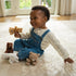 GUND Kai plush teddy bear in light beige soft fabric with rounded shape, small stuffed toy held by smiling child seated on carpet with other plush animals in a bright home setting