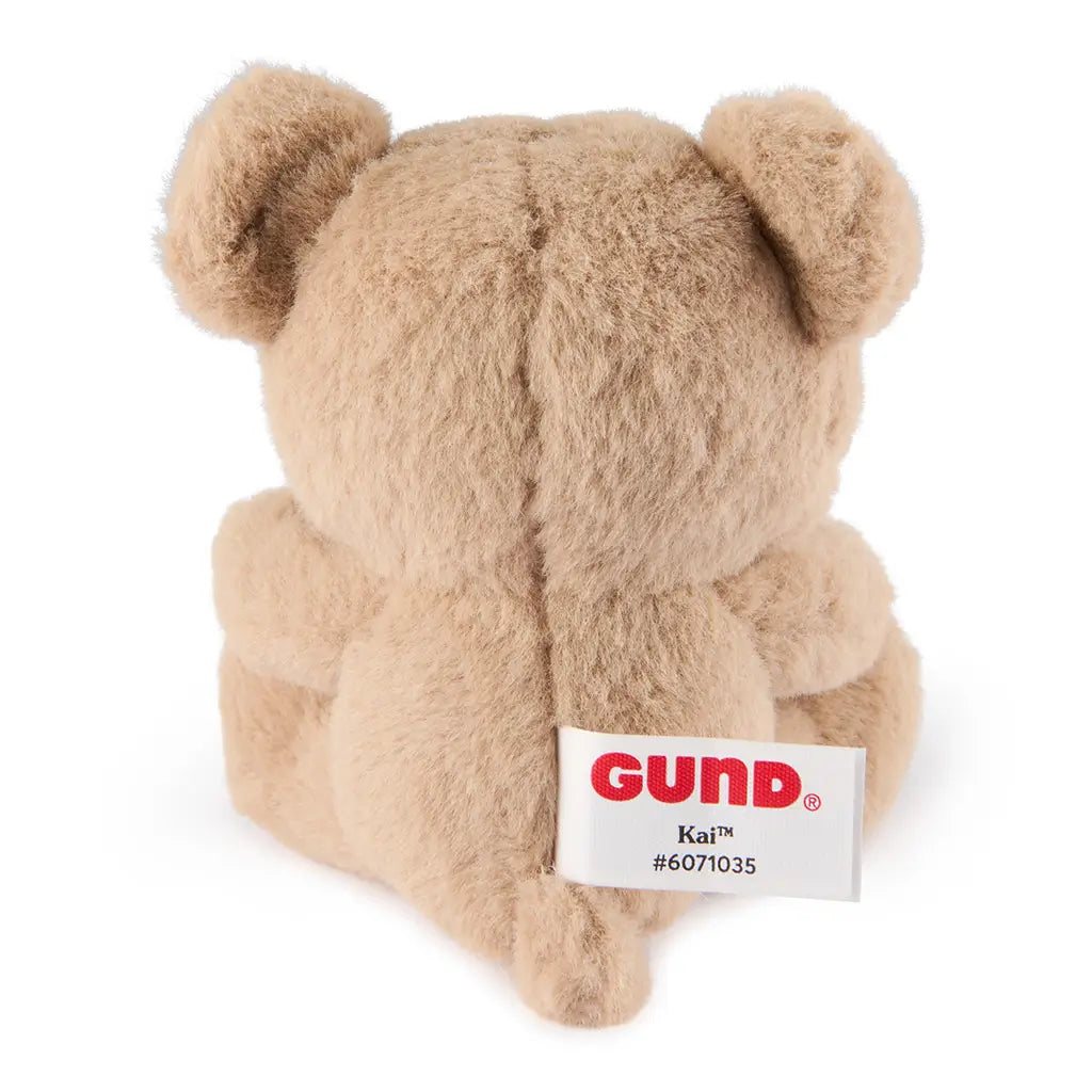 GUND Kai plush teddy bear in light beige soft fabric with rounded compact shape, rear view showing seam detail and brand tag, small cuddly toy displayed on a white background