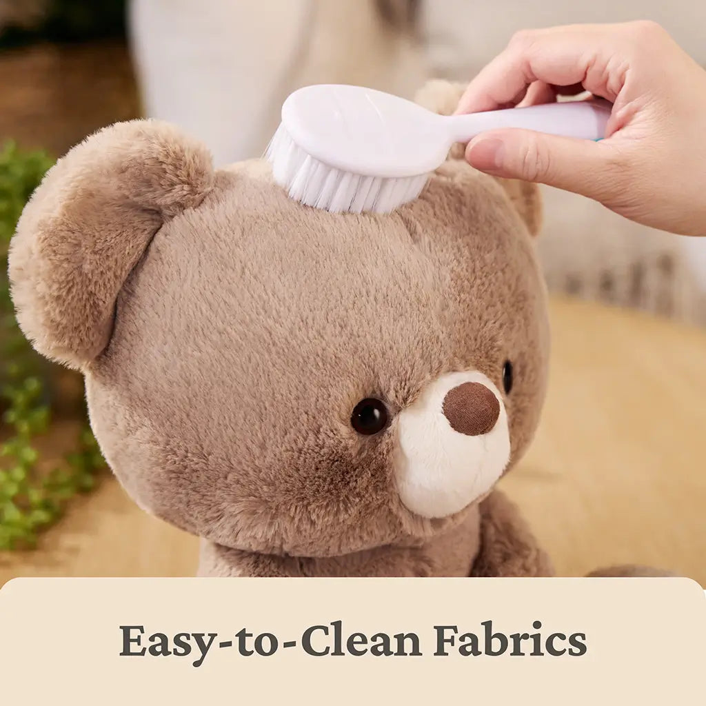 GUND teddy bear easy-clean feature image showing warm beige plush fur, ultra-soft textured fabric, rounded bear head, being gently brushed with a white handheld brush to highlight washable, easy-care materials for everyday use.
