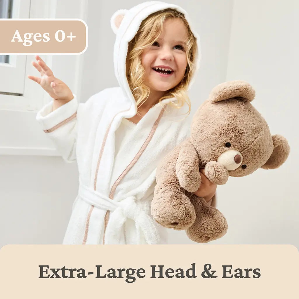 GUND teddy bear lifestyle image showing beige plush fur, oversized rounded head and ears, and a soft squishy body, held by a smiling child in a bright indoor setting to illustrate scale, comfort, and suitability for all ages.