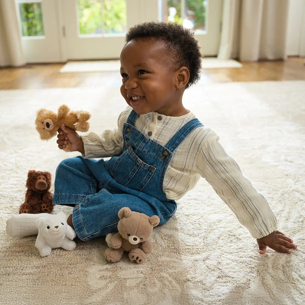 GUND Toothpick plush teddy bear in light tan fuzzy fabric with soft rounded shape, small size toy held by smiling child, surrounded by stuffed animals on carpet in bright home living room setting