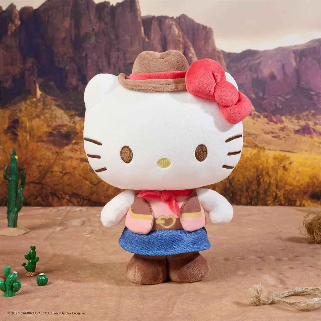 Hello Kitty cowgirl plush by GUND in white velour fabric with soft texture, rounded standing form, embroidered facial details, brown cowboy hat with red band, pink western outfit, and blue skirt, staged outdoors in a desert canyon scene.