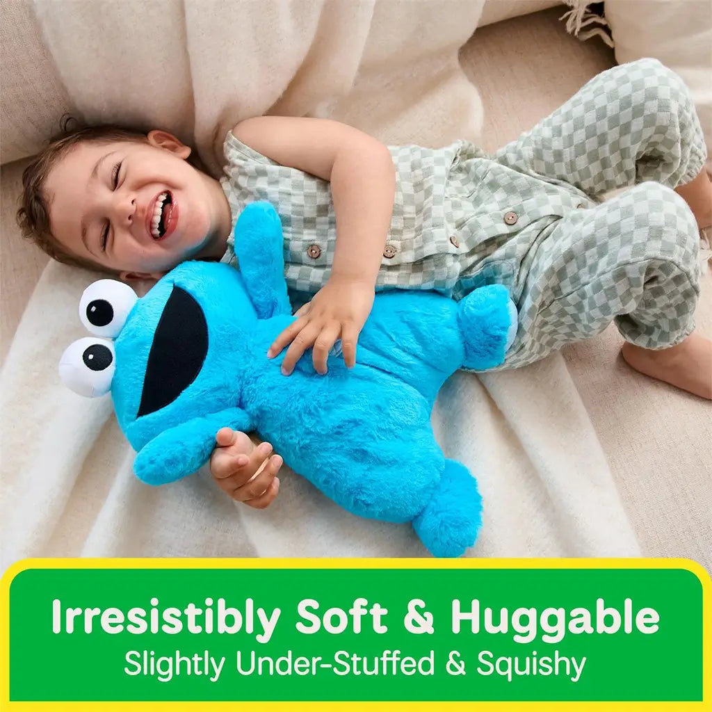 GUND Sesame Street Cookie Monster plush in bright blue plush fabric with floppy arms and slightly under-stuffed body, designed for melt-in-your-arms comfort, shown cuddled by a laughing toddler on a sofa.