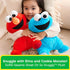 GUND Sesame Street Elmo and Cookie Monster plush toys in bright red and blue fabrics with rounded huggable forms, designed for comforting play, held closely by a child seated indoors in a cozy living room setting.