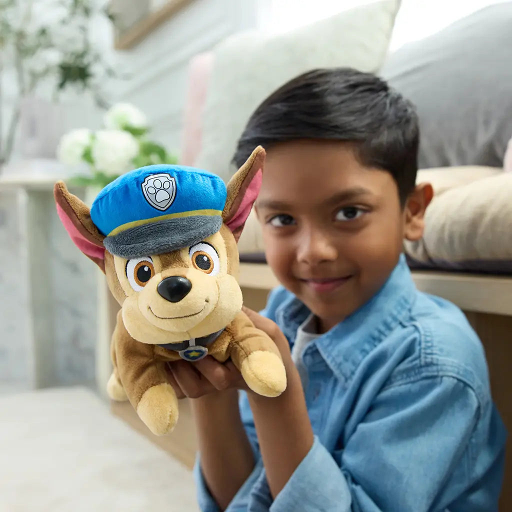 Paw Patrol Chase plush toy in brown and tan soft fabric with blue police hat, small rounded dog form held by smiling child in a bright indoor living room environment